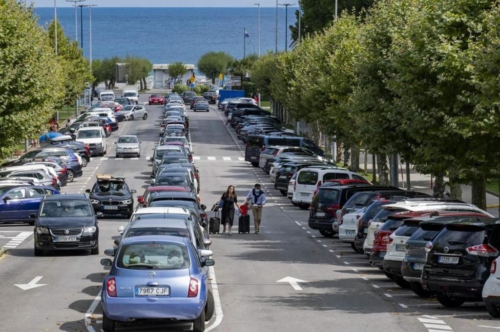 Coches aparcados a ambos lados de la carretera, árboles a los lados. Gente caminando con equipaje. El océano al fondo.