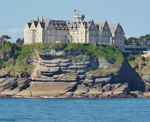 Palacio en lo alto de un acantilado con vistas al océano; arquitectura de piedra, cielo azul claro y agua.