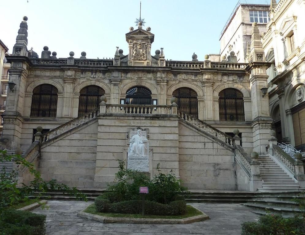 Edificio de piedra con escaleras, ventanas arqueadas y una estatua en un patio.