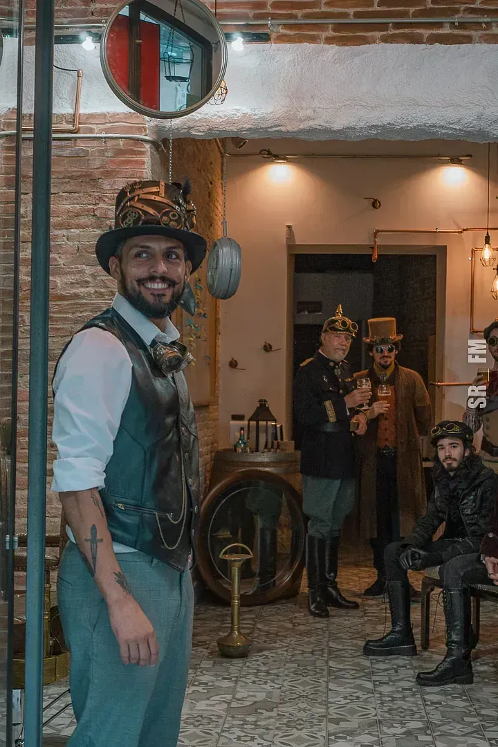 Un hombre vestido con traje steampunk sonríe y posa al aire libre; otros disfrazados en el fondo.