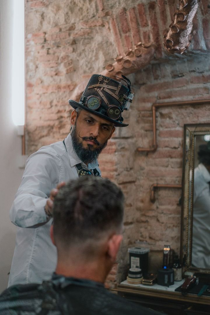 Barbero con sombrero de copa steampunk haciendo un corte de pelo frente a una pared de ladrillos.