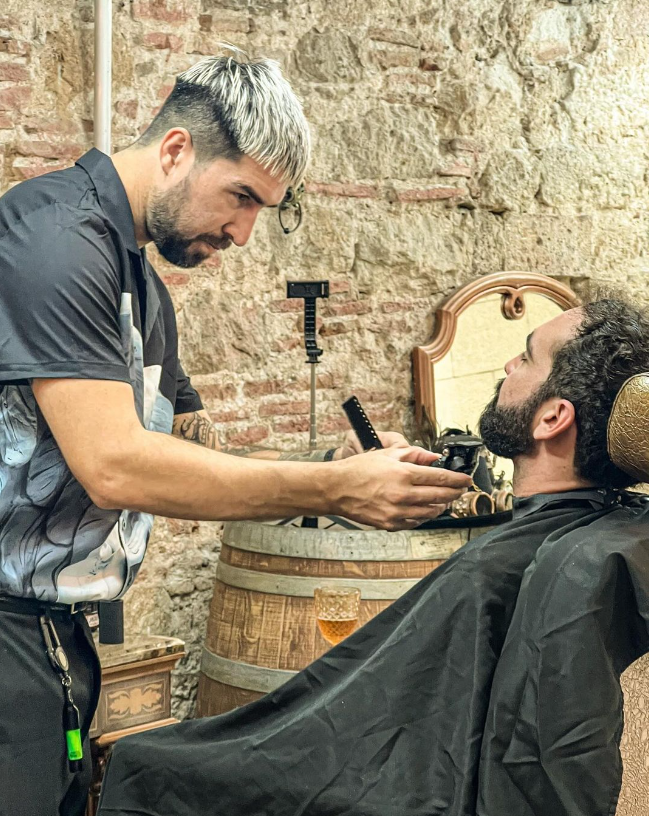 Un barbero recorta la barba de un cliente en un local rústico. El barbero tiene el pelo claro y una expresión concentrada.