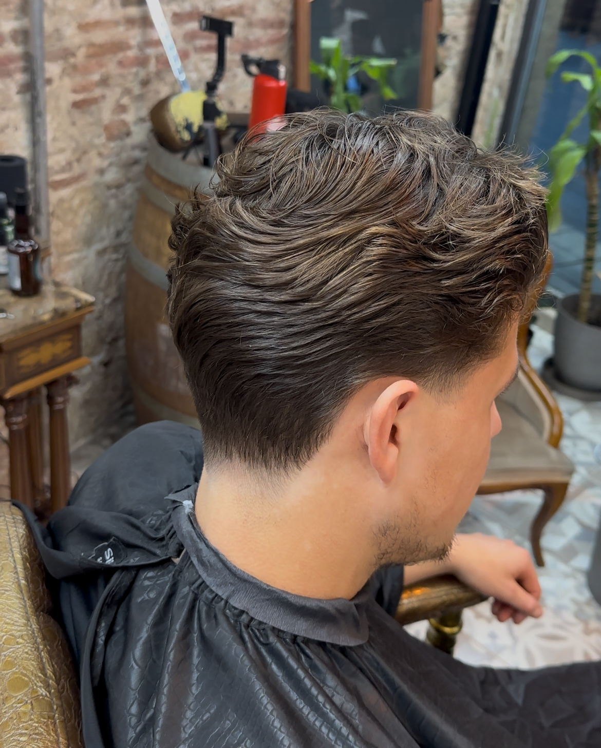 Hombre con cabello castaño texturizado y los laterales descoloridos. Sentado en una silla de barbero, en un interior.