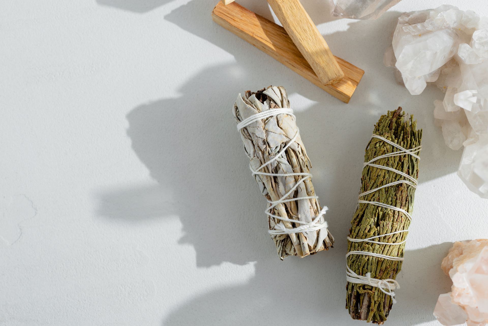 Des fagots de sauge blanche et verte, des bâtonnets de Palo Santo et des cristaux de quartz sur une surface blanche.
