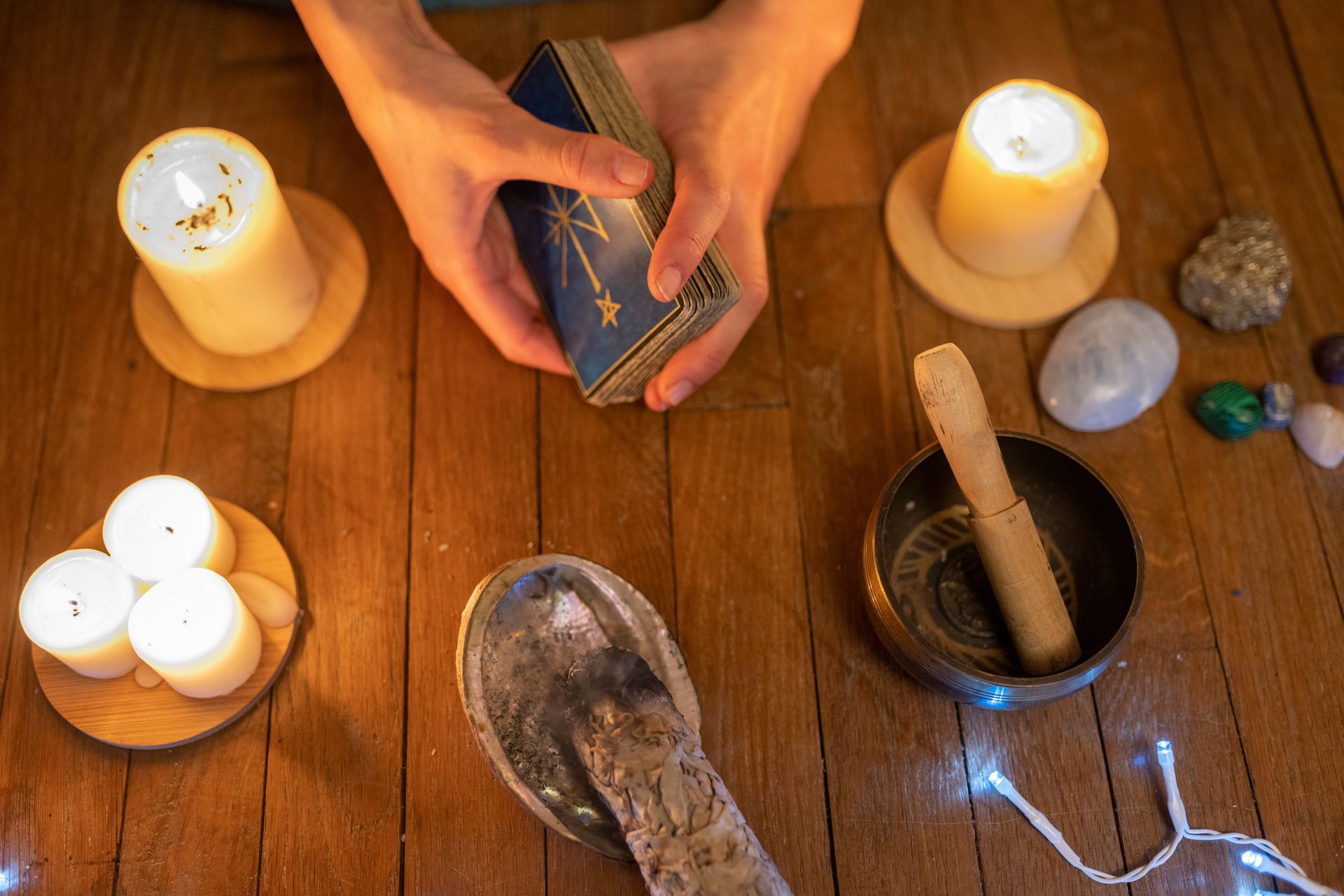 Des mains tenant un jeu de tarot, des bougies, des pierres et un bol chantant sur une table en bois.