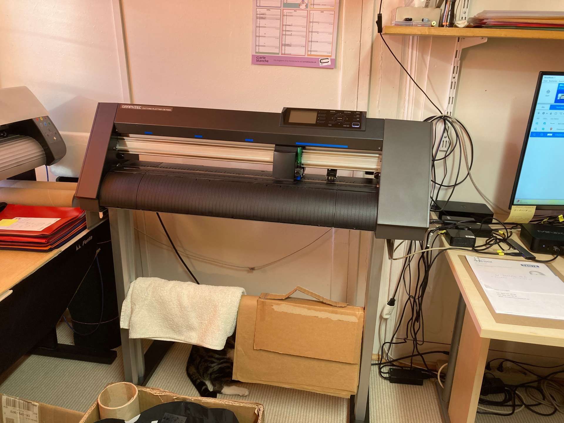 Ein Vinylschneider mit Bedienfeld steht auf einem Metallständer in einem unordentlichen Arbeitsbereich.