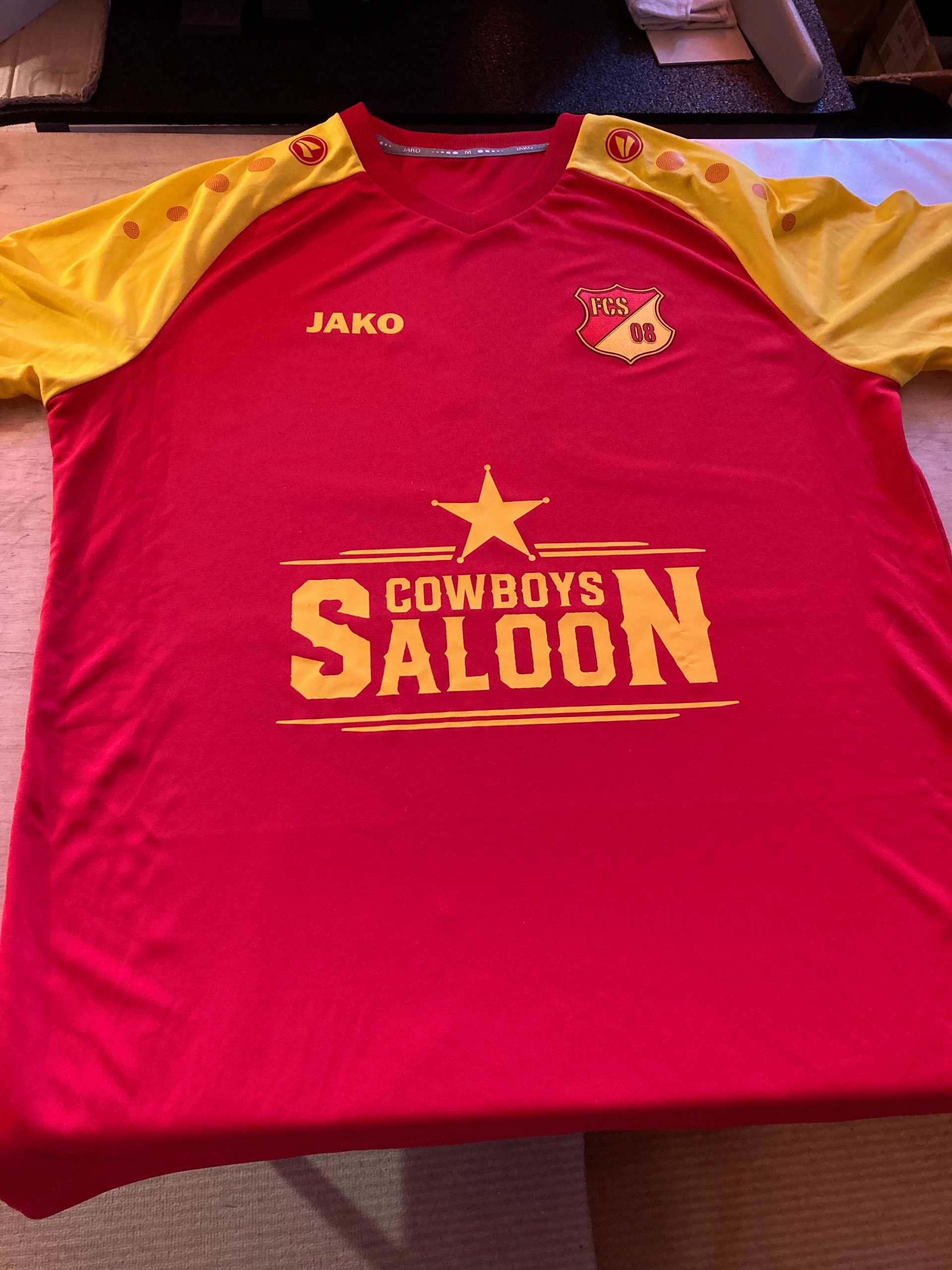 Rot-gelbes Fussballtrikot mit dem Logo „Cowboys Saloon“.