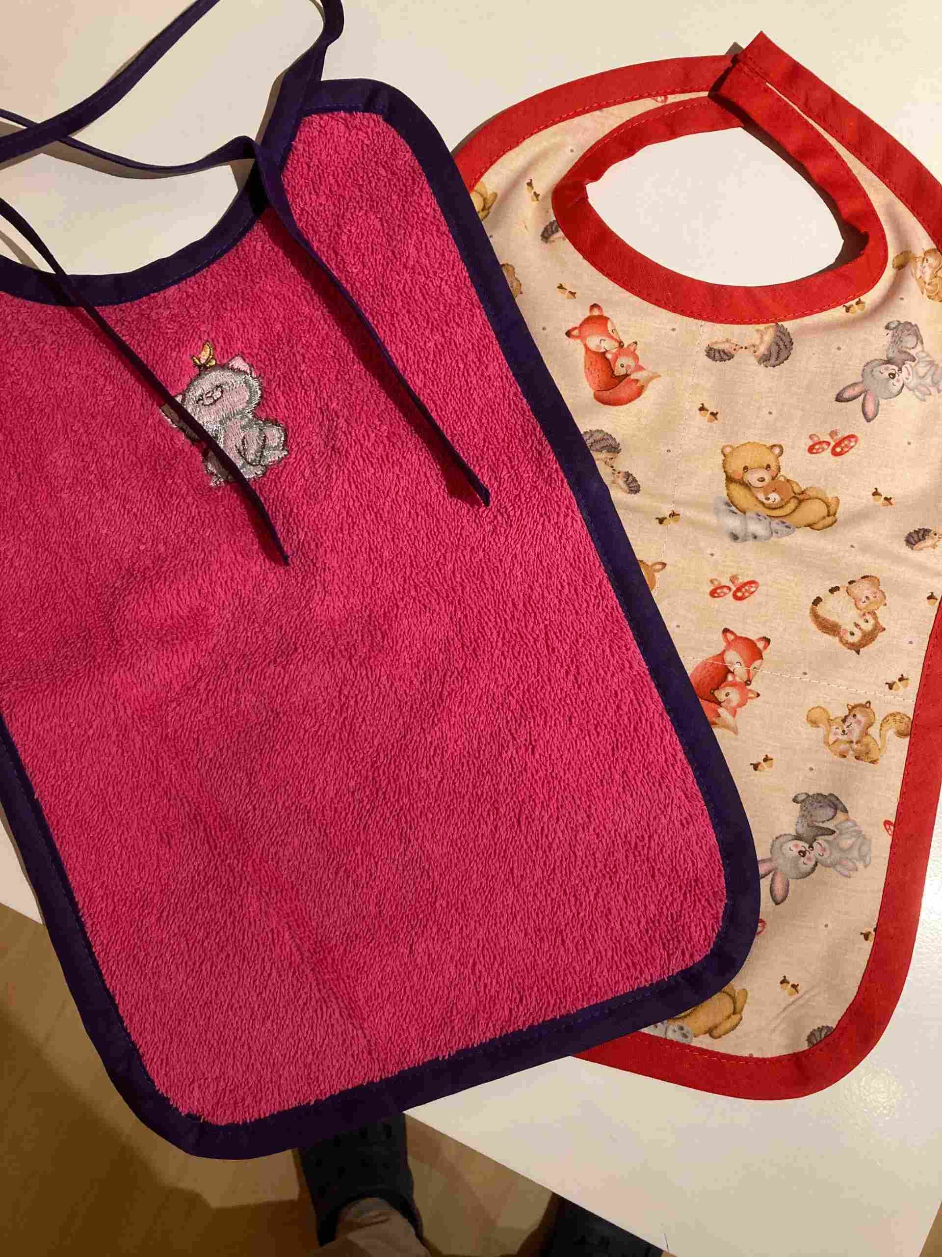 Zwei Baby-Lätzchen: ein rosa Lätzchen mit grauem Rand und Bärenmotiv; ein gemustertes Lätzchen mit rotem Rand.