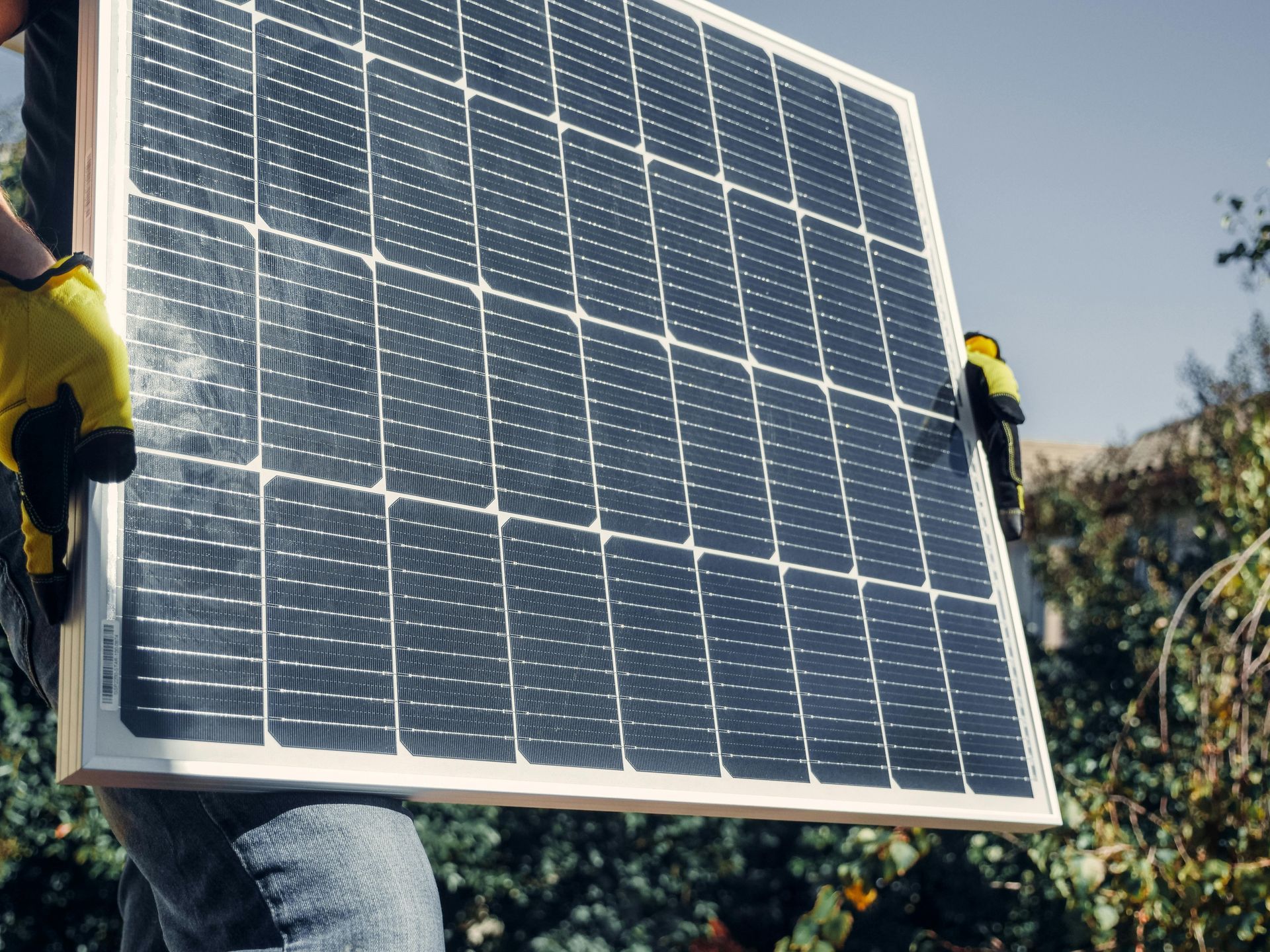 Una persona con guantes de trabajo amarillos transporta un panel solar al aire libre, frente a una vegetación verde.