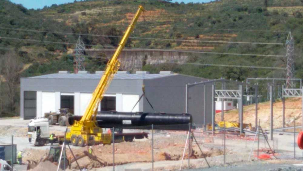 Una gran grúa amarilla está levantando una tubería grande frente a un edificio.