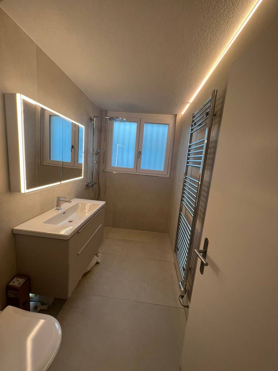 Modernes Badezimmer mit Waschtisch, Spiegel, Fenster und Handtuchwärmer, beleuchtet durch LED-Deckenleuchten