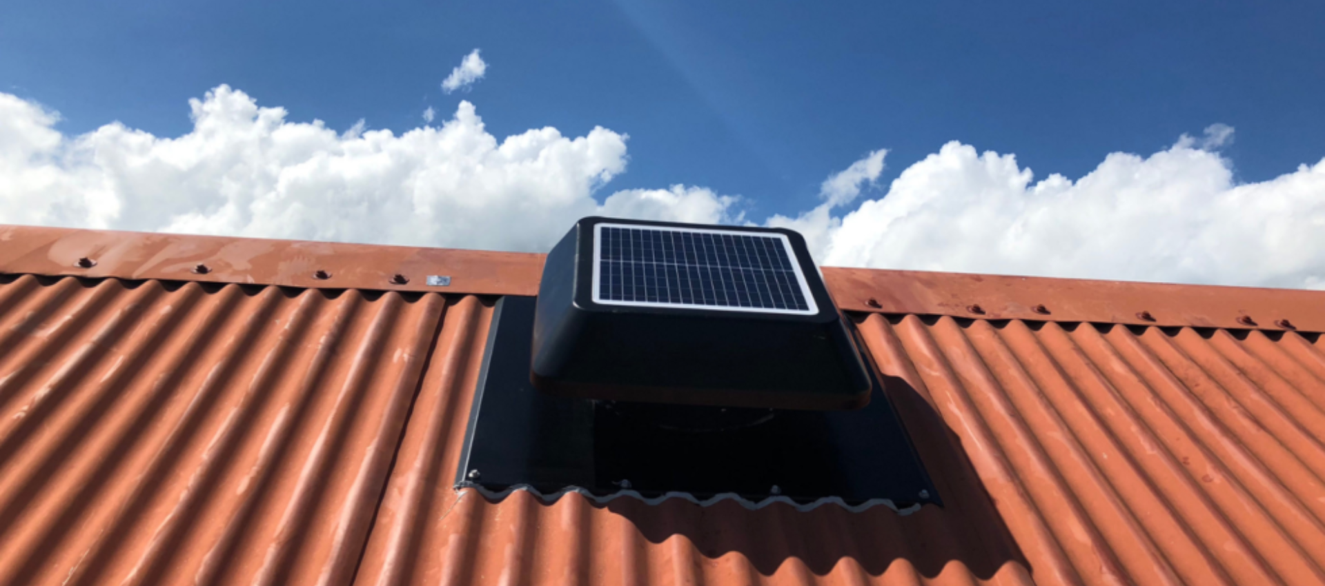 Aérateur de toit à énergie solaire sur un toit en tôle ondulée rouge, sur fond de ciel bleu nuageux.