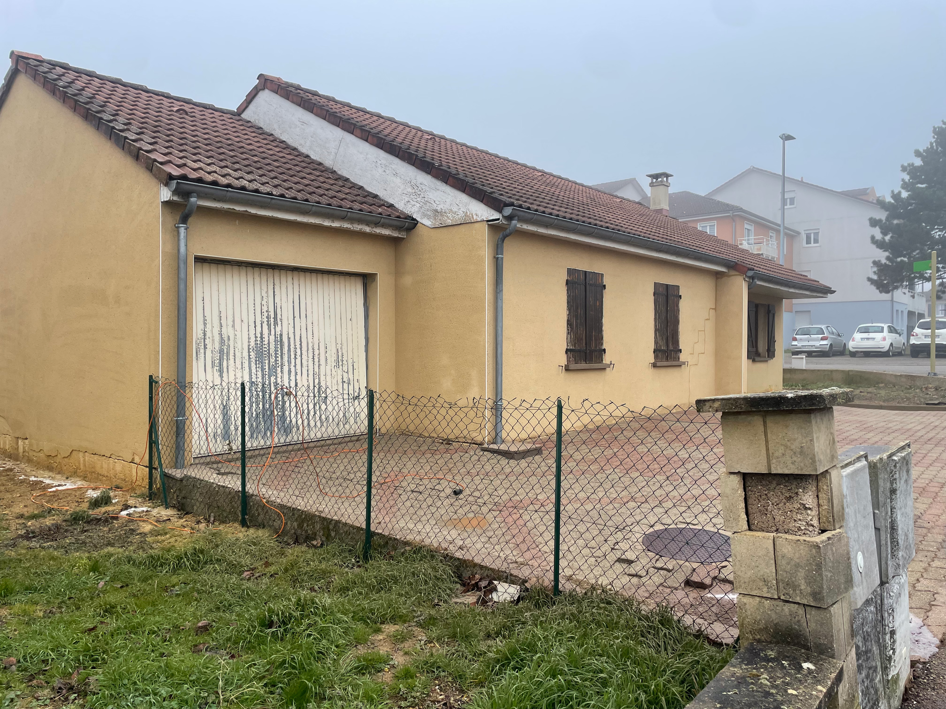 Bâtiment jaune avec porte de garage et cour clôturée ; temps couvert.