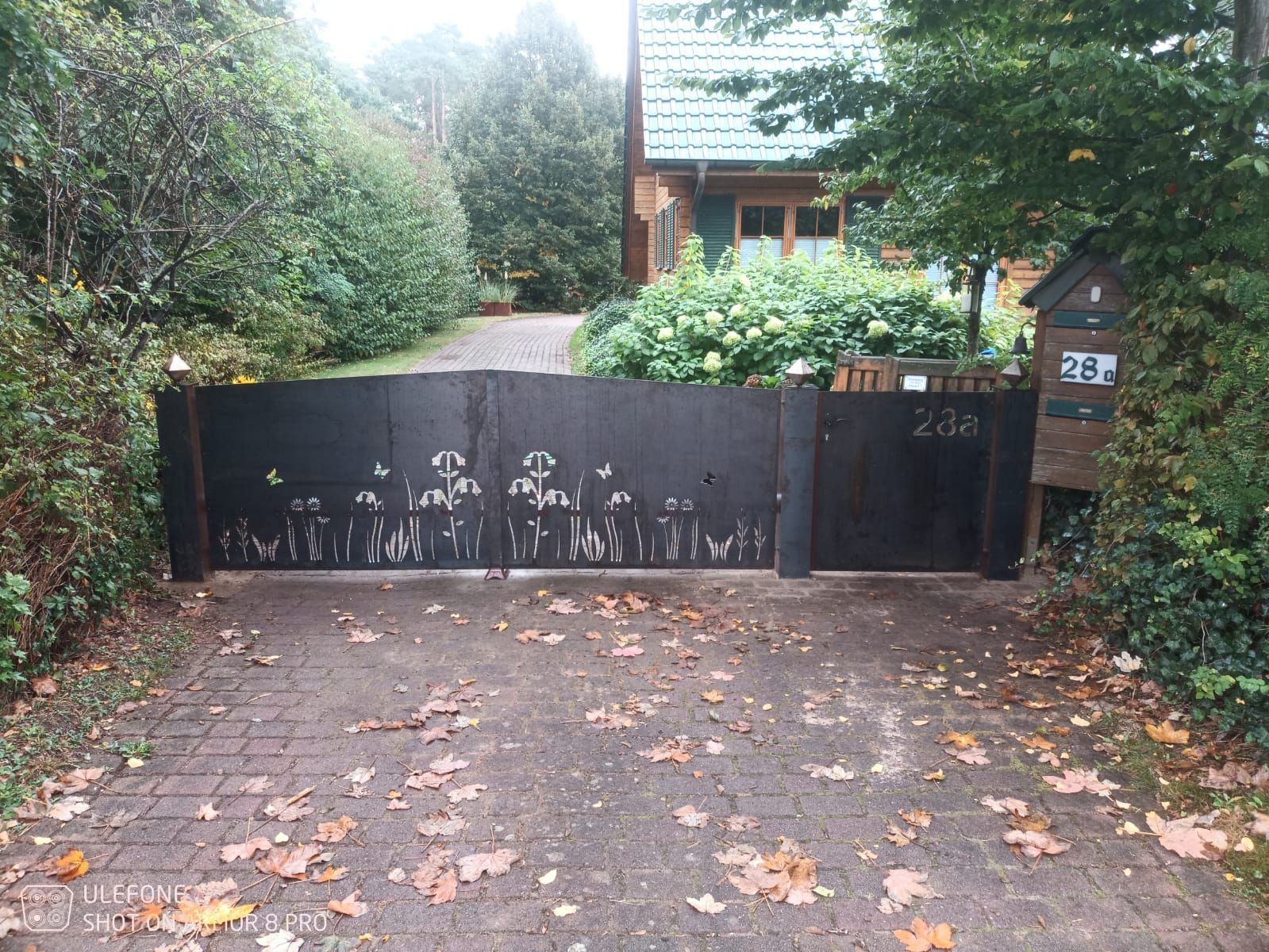 Ein schwarzes Tor versperrt eine von Grün umgebene Einfahrt; dahinter ist ein Haus zu sehen.