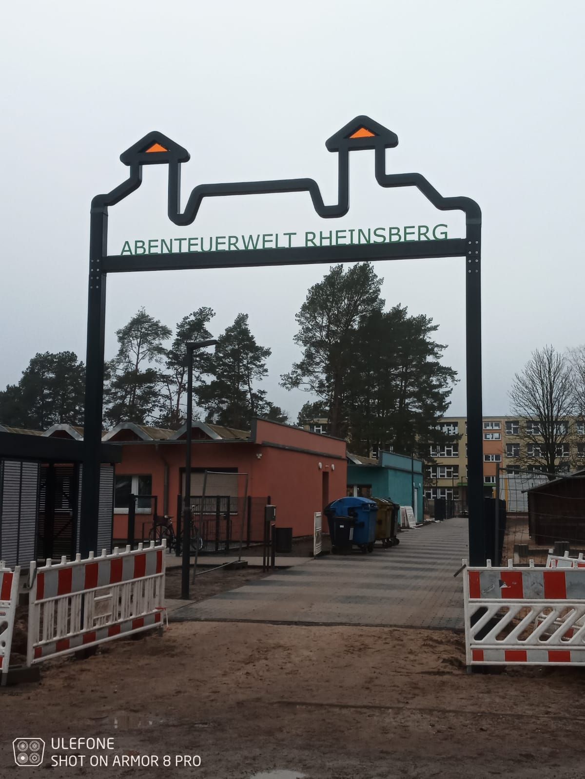 Eingangstor mit dem Schild „Abenteuerwelt Rheinsberg“, Absperrungen und einem unbefestigten Parkplatz unter bewölktem Himmel