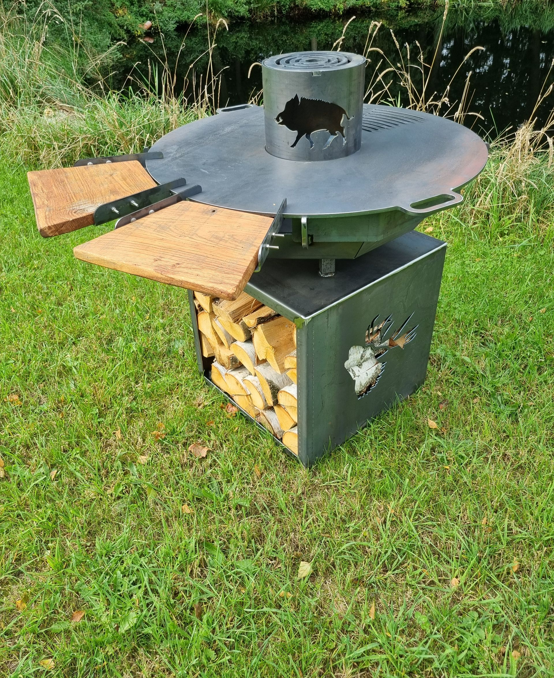 Im Freien steht eine Feuerstelle aus Metall mit runder Oberseite und seitlichen Ablagen, daneben gestapeltes Brennholz.