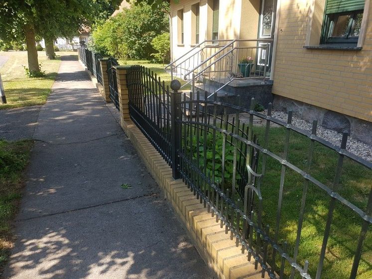 Gehweg neben einem gelben Gebäude mit schwarzem Zaun und kleiner Eingangstreppe in einem grünen Wohngebiet