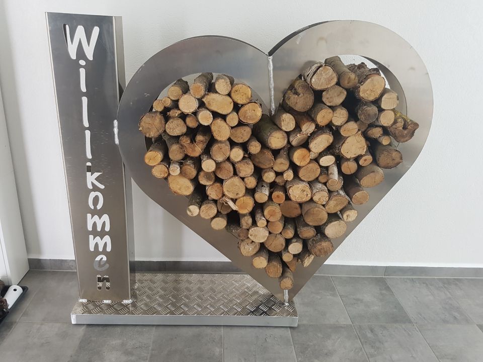 Herzförmiger Holzständer mit gestapeltem Brennholz und einem senkrechten „Willkommen“-Schild auf grauem Fliesenboden