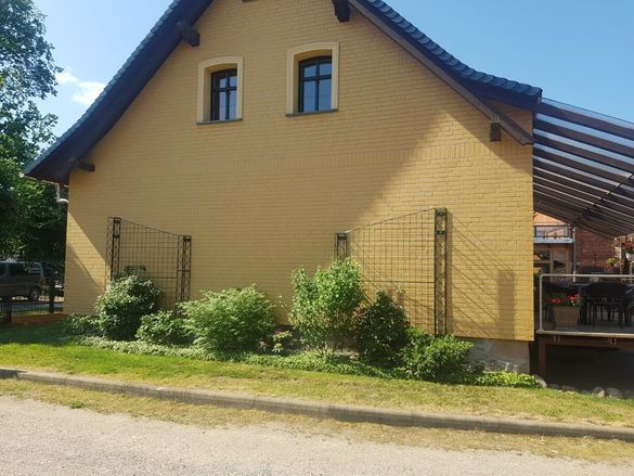 Ein gelbes Backsteinhaus mit steilem, dunklem Dach, kleinen Fenstern und Gartensträuchern an einer Straßenseite