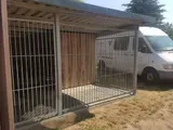 Kleiner beigefarbener Hundezwinger mit weißen Metallstäben neben einem weißen Lieferwagen in einem Hof