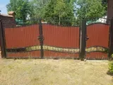 Rotes Metalltor mit Backsteinsäulen und dahinter grünen Bäumen, über trockenem Gras davor