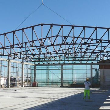 Almacén con estructura de acero en construcción con trabajadores sobre una losa de hormigón bajo un cielo azul despejado.
