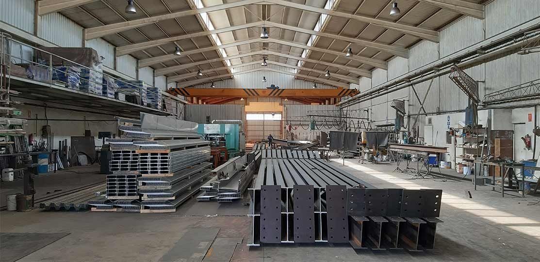Gran nave industrial con vigas metálicas apiladas y maquinaria bajo un techo alto.