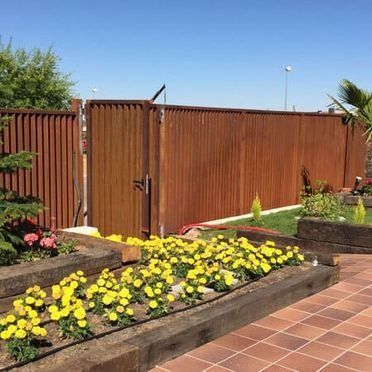 Una verja metálica color óxido detrás de macizos de flores con flores amarillas y rosas junto a un patio embaldosado bajo un cielo azul.