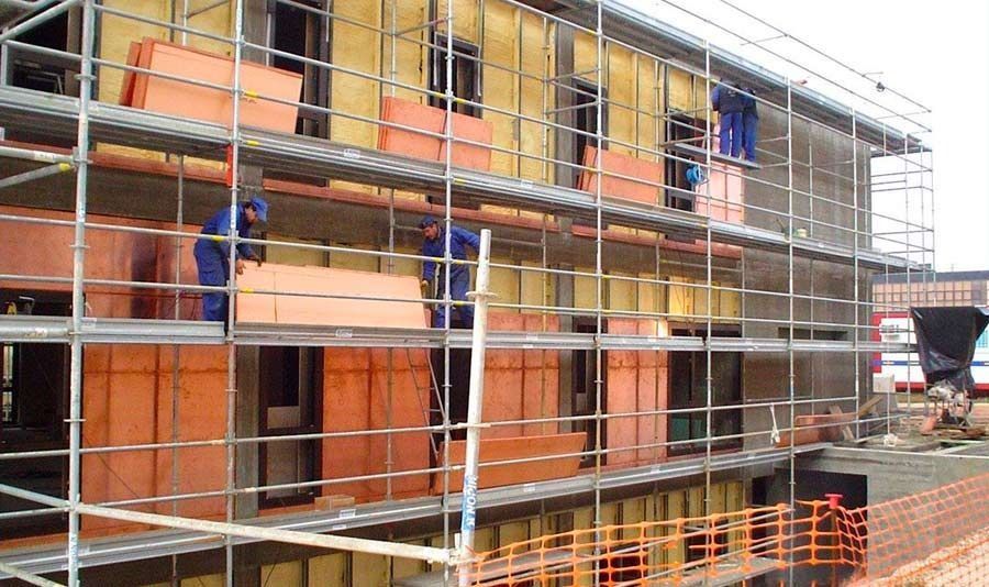 Andamios alrededor de un edificio en construcción con paneles naranjas y amarillos y trabajadores al fondo.