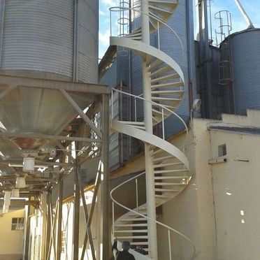 Escalera blanca en espiral junto a grandes silos industriales de grano y estructuras de soporte metálicas.