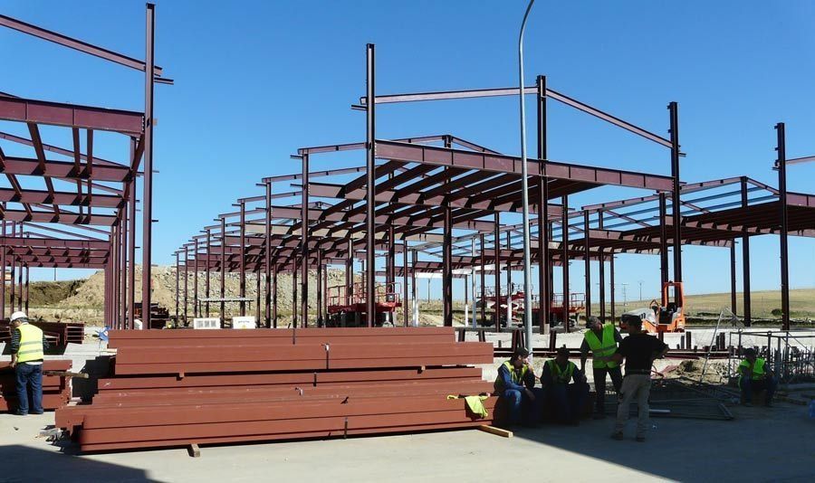 Obra en construcción con estructura de acero, vigas apiladas y trabajadores con chalecos de seguridad.