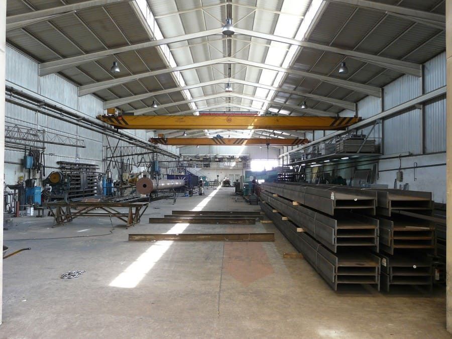 Interior de un taller de fabricación de metales con vigas de acero apiladas y una grúa puente amarilla.