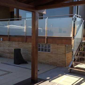 Terraza en la azotea con barandilla de cristal, postes de madera, escaleras y una vista difusa de la ciudad al fondo.