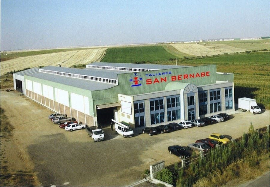 Vista aérea de un gran edificio industrial con coches aparcados, rodeado de campos abiertos y con letreros de SAN BERNARD.