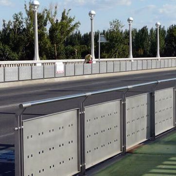 Pasarela junto a un puente de carretera con barreras metálicas, farolas y árboles más allá de la barandilla.
