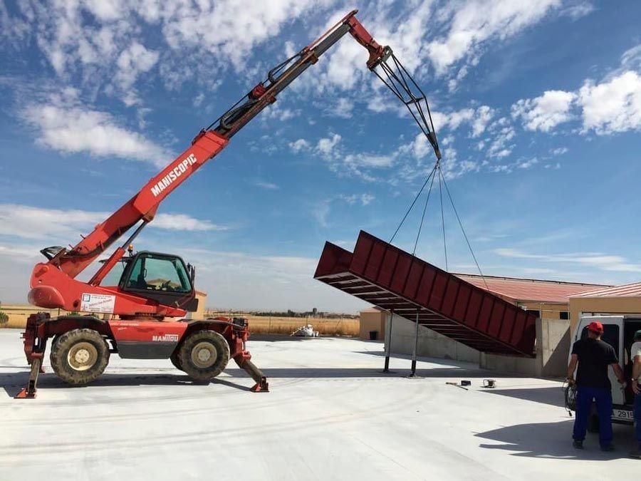 Una grúa roja levanta un gran contenedor de basura metálico y lo coloca en su lugar en una azotea bajo un cielo azul.