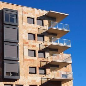 Edificio de apartamentos moderno con fachada de piedra beige y balcones de cristal apilados contra un cielo azul.