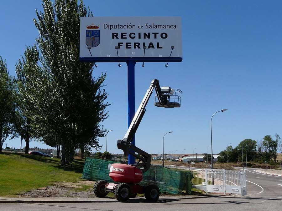 Una grúa elevadora está subiendo a un trabajador junto a un letrero que dice “Recinto Ferial” al costado de la carretera.