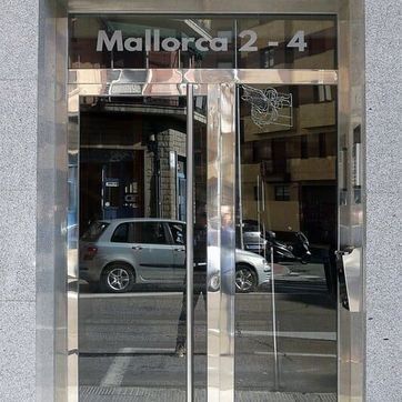 Entrada de cristal con letrero de “Mallorca 2-4” y coches aparcados reflejados en las puertas.