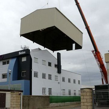 Un gran módulo prefabricado está siendo izado por una grúa sobre un edificio industrial blanco y azul.