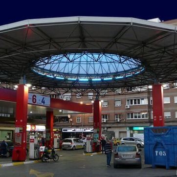 Marquesina de gasolinera con coches, surtidores y un contenedor azul de reciclaje en un entorno urbano.