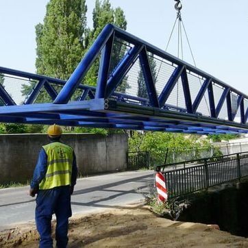 Una grúa eleva la estructura azul de un puente sobre una carretera, con un trabajador con casco amarillo debajo.