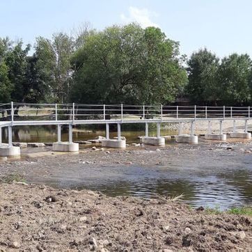 Puente peatonal de hormigón sobre un río rocoso poco profundo con árboles al fondo.