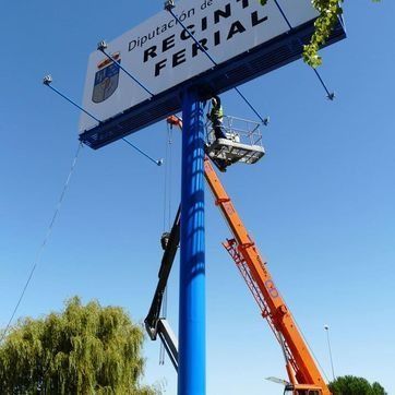 Trabajadores subidos a una plataforma elevadora naranja instalan un gran letrero en la parte superior de un poste azul al borde de la carretera, bajo un cielo despejado.