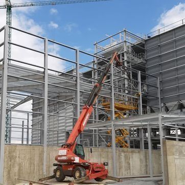 Una carretilla elevadora telescópica roja levanta vigas de acero dentro de un edificio en construcción.