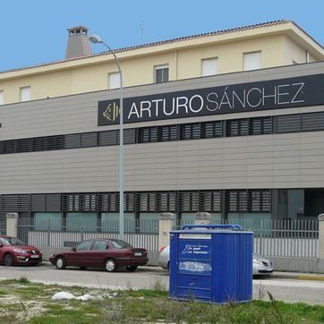 Edificio de la fábrica ARTUROSÁNCHEZ con coches aparcados y un contenedor de basura azul delante.