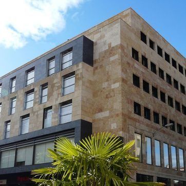 Edificio de oficinas moderno de varias plantas con fachada de piedra color tostado, secciones de ventanas oscuras y una palmera en la fachada.