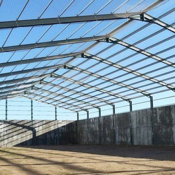 Estructura vacía similar a un almacén, con techo de armazón metálico, paredes de hormigón y un cielo azul brillante en lo alto.