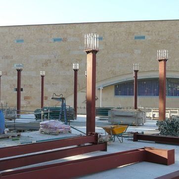 Obra en construcción con vigas de acero, columnas rojas y materiales de construcción frente a un muro de ladrillos.