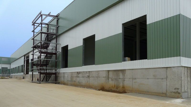 Exterior de una nave industrial con escaleras metálicas y revestimiento verde y blanco sobre una base de hormigón.